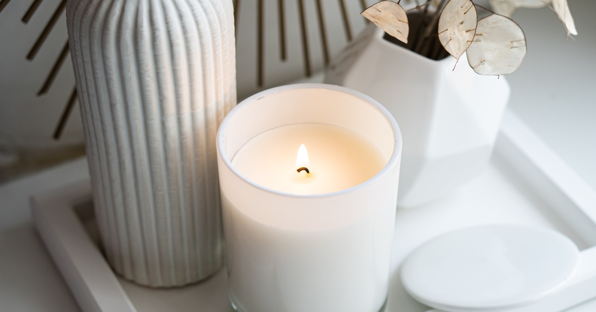 A candle sitting on a tray next to a bar of soap, a small potted plant, and a thin vase. All of these accessories are white.