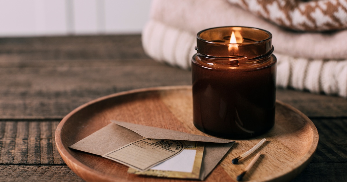 A small candle sitting on a wooden tray next to a brown envelope and some used matches. There are some blankets behind it.