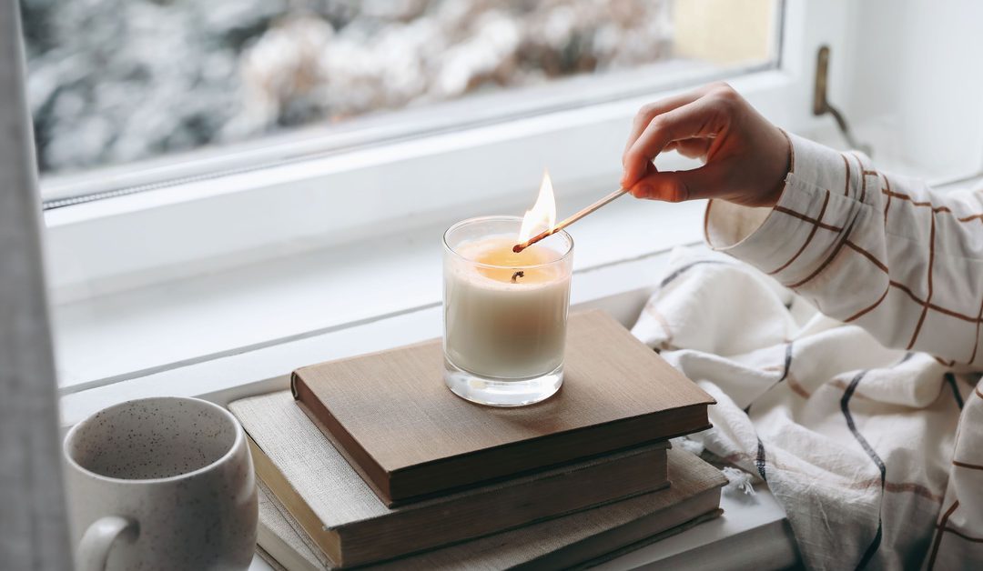 Someone lighting a candle with an extra-long match. The candle is sitting on some books next to a windowsill.