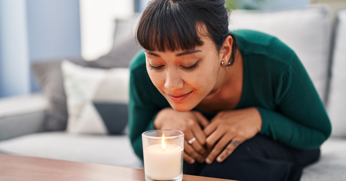 A young woman sitting on a sofa while smelling an aromatic candle in a bright and comfortable home interior.