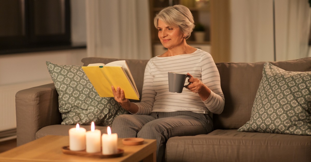 A senior woman reading a book while holding a mug at home in the evening, with 3 candles lit in front of her.
