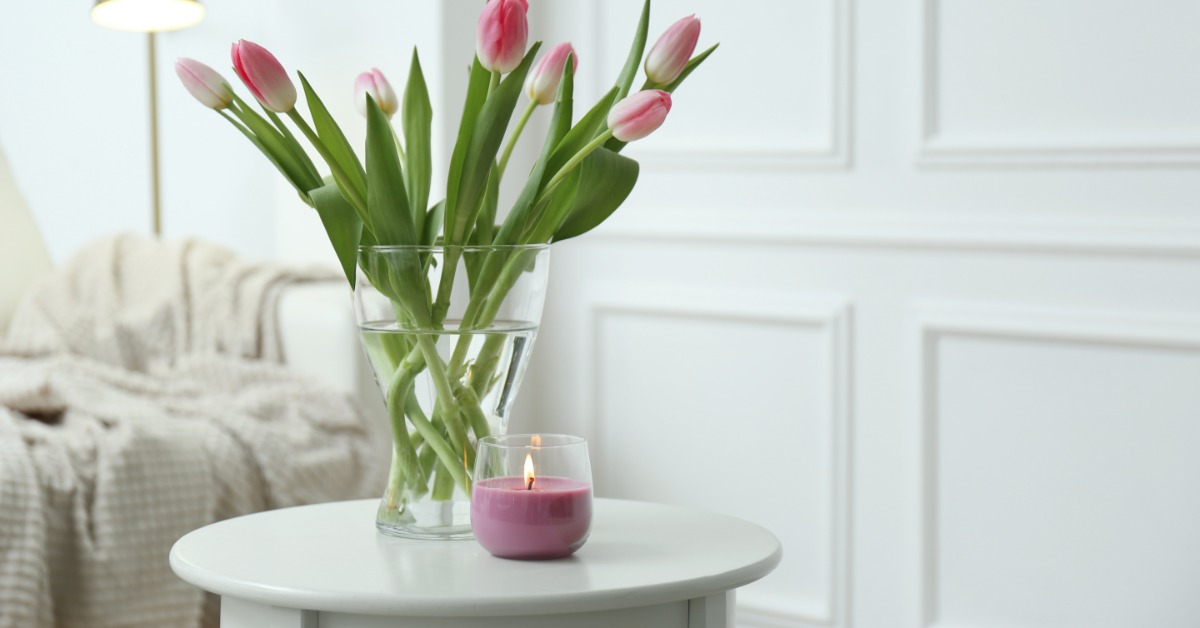 Pink tulips and a burning candle arranged on a white table indoors, with a couch and a floor lamp further behind it.