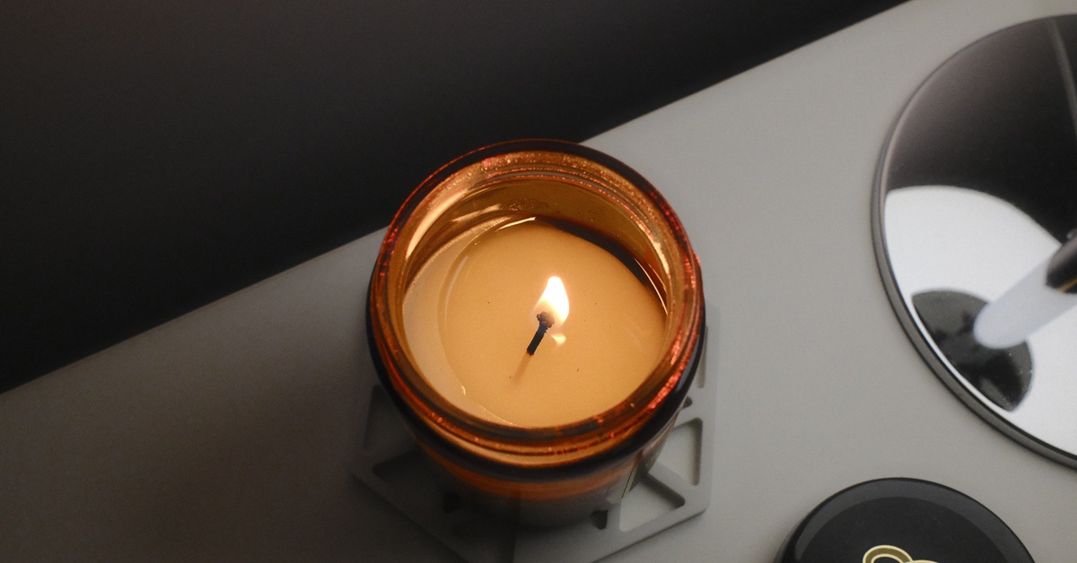An overhead view of a lit candle burning inside an amber glass container on a white coaster that's sitting on an end table.