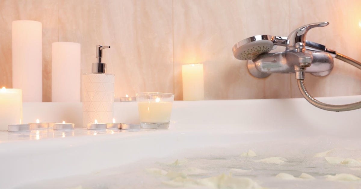 A bathtub with floating flower petals and bubbles. There are quite a few burning candles next to a bottle of lotion.