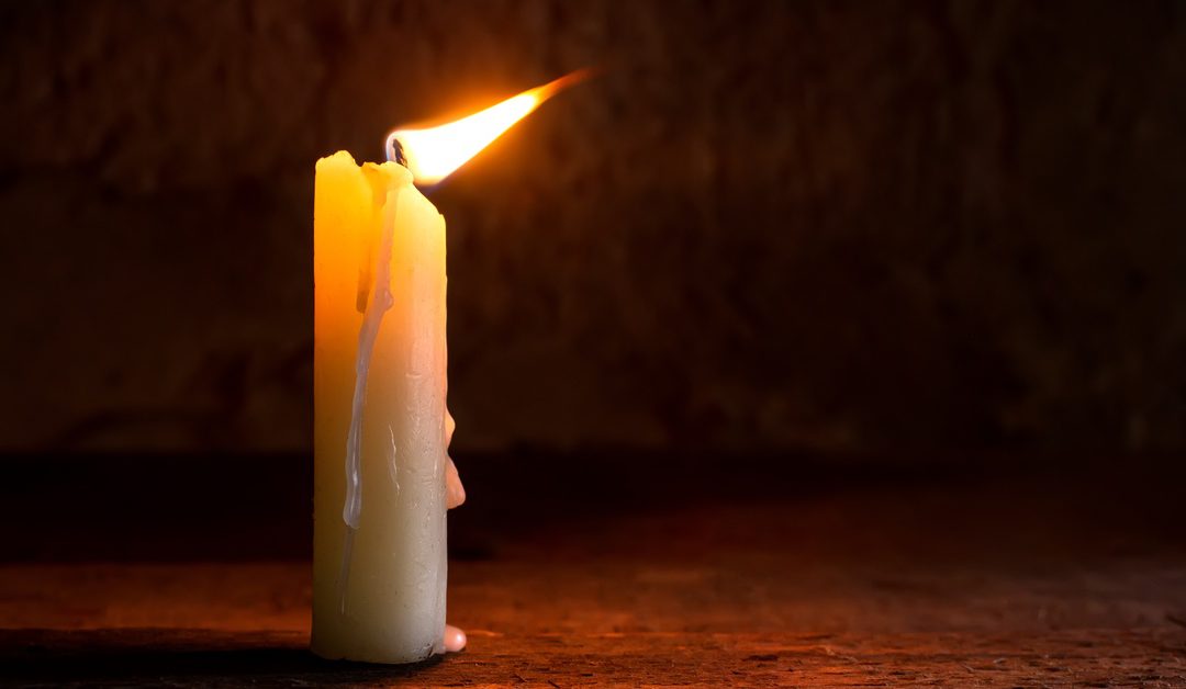 A lit candle burning on a wooden surface while its flame moves with the wind. The candle is also dripping hot wax.