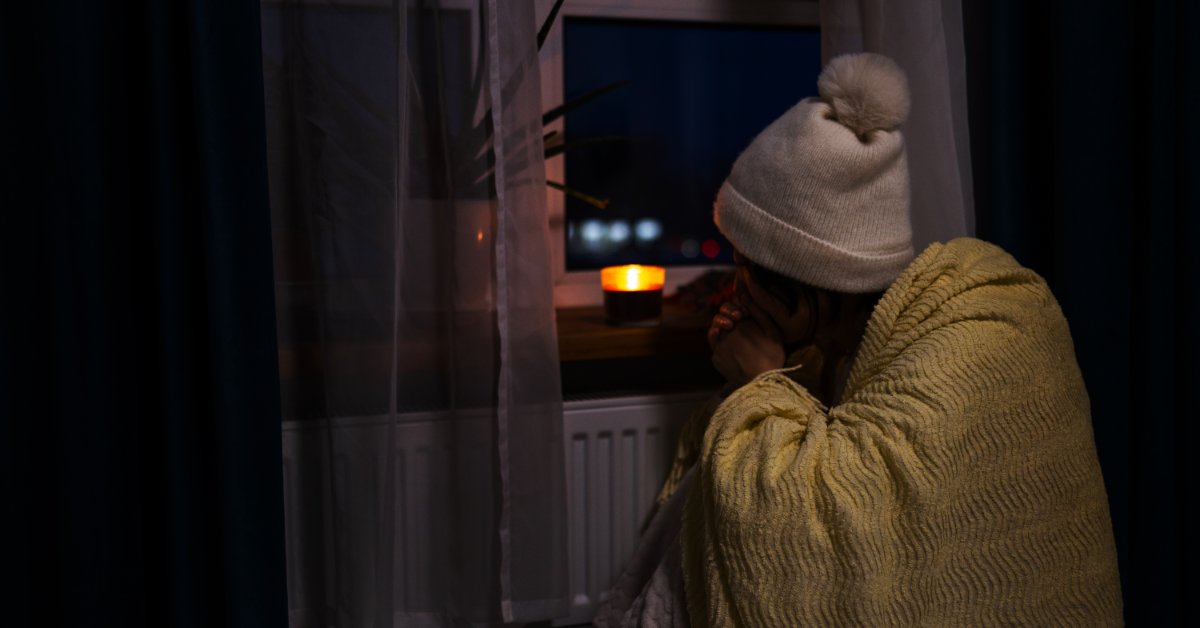 A girl sitting near a window in a dark room, next to a small burning candle that lights the surrounding space.