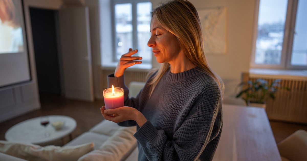 A smiling woman holding a burning candle in a glass container while walking through the living room of her apartment.