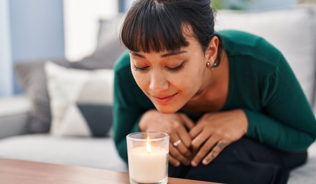 A woman sitting on a sofa while leaning forward to smell a candle sitting on the edge of her coffee table.