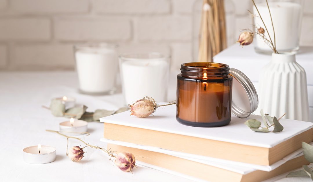 A burning candle sitting on some white books with eucalyptus leaves and dried flowers arranged around it.