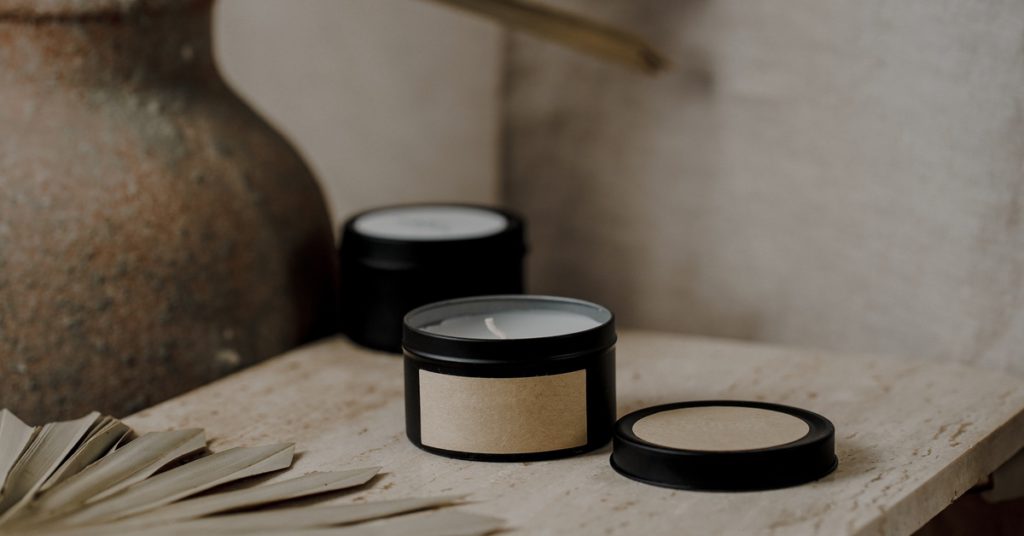 A black candle jar with a beige label sitting on a worn wooden table beside some dried leaves and a clay vase.