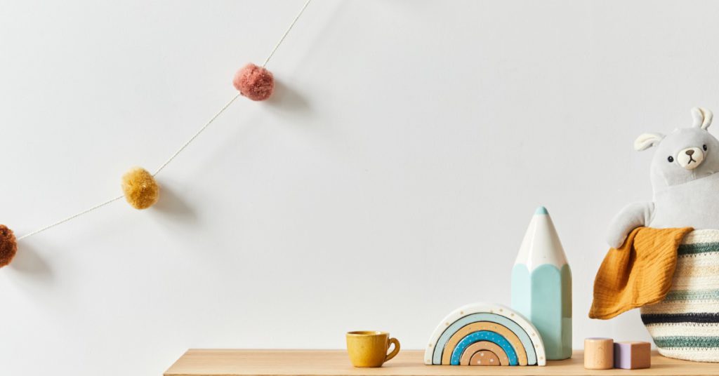 A baby room with a teddy bear, toys, and a coffee cup, with a string of cotton balls hanging against a white wall.