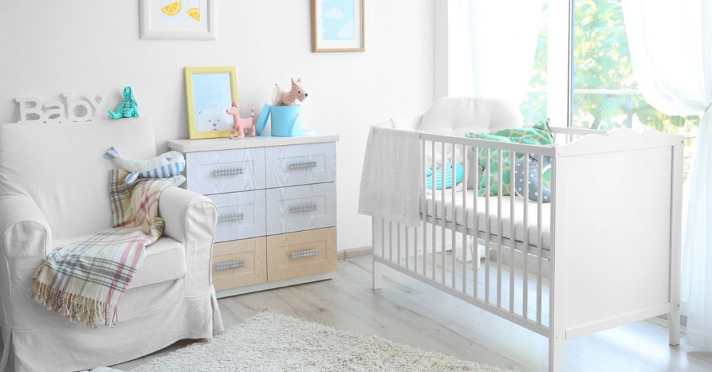 A modern baby room with a crib, armchair, rug, and soft decor arranged beside a window with light coming in.