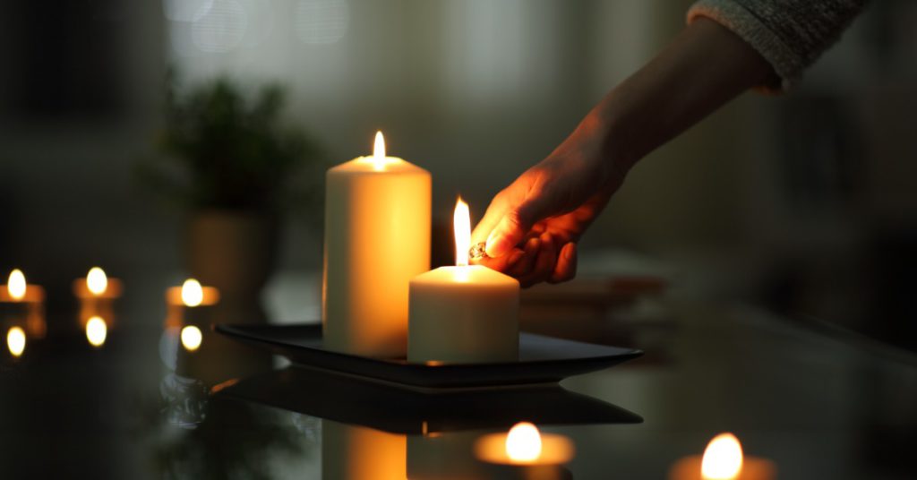 A hand lighting a candle with a lighter in a dark room, with a warm flame illuminating the candle top.