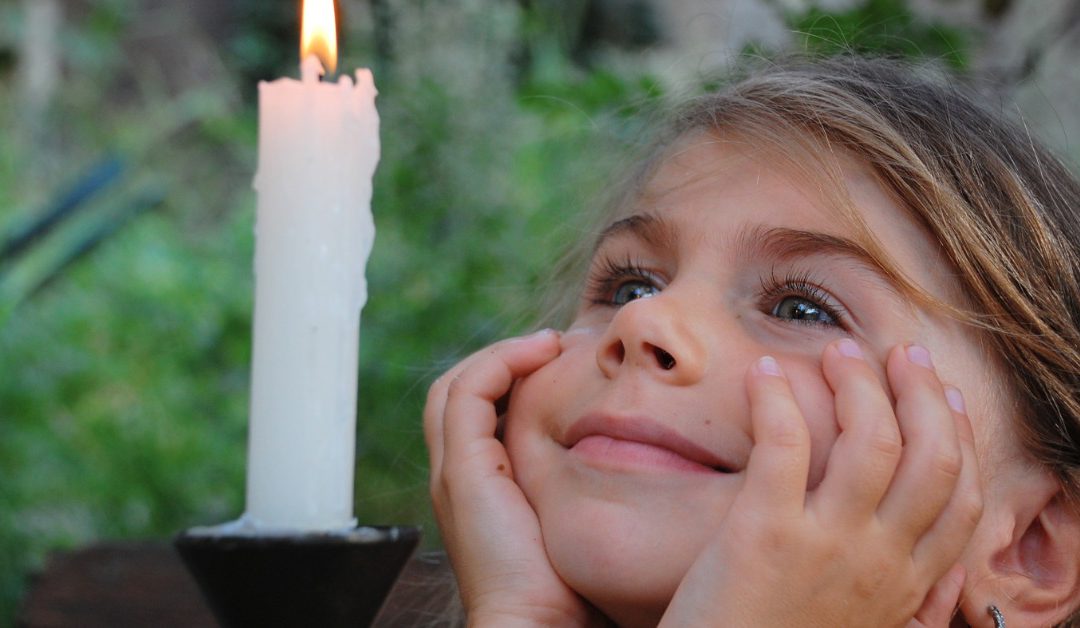 A girl resting her face in her hands while looking toward a lit white candle with some greenery behind her.
