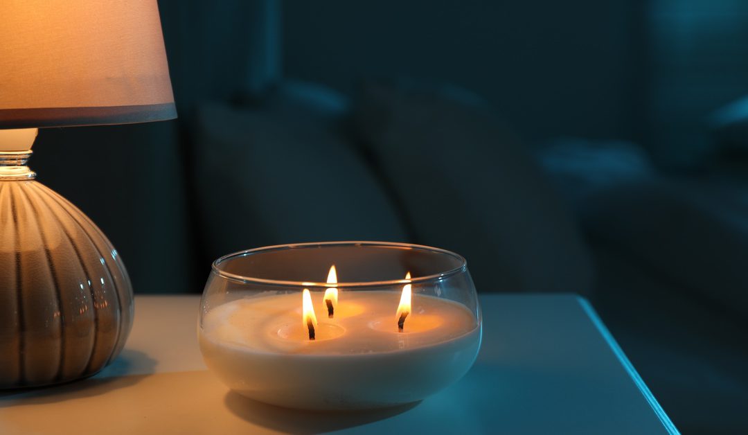 A glass candle with three flames sitting on a bedside table beside a glowing lamp in a dark bedroom.