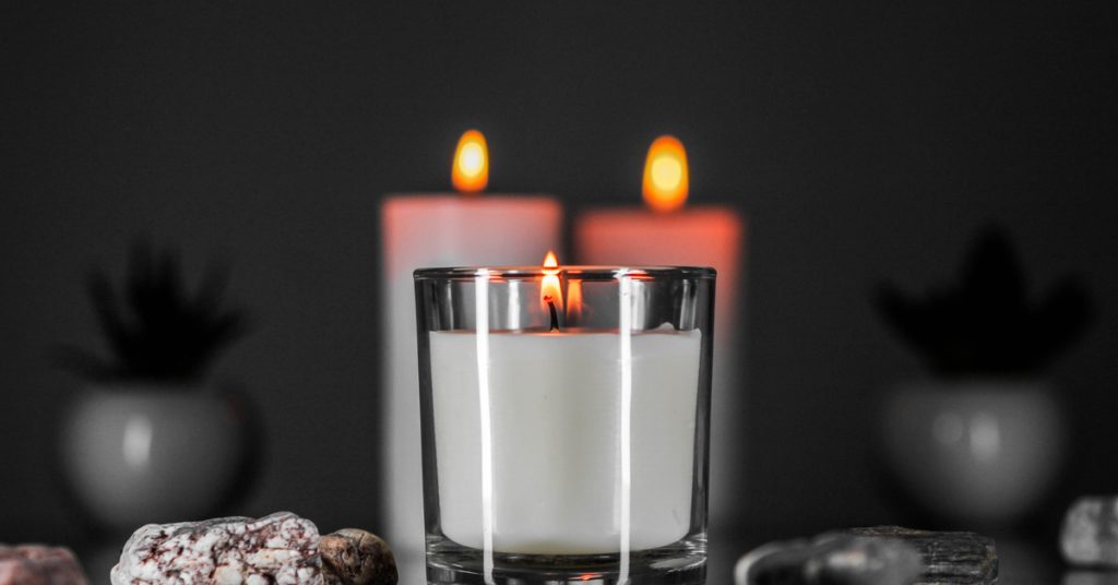A lit glass candle sitting on a table with stones next to it. There are two other lit candles further behind it.