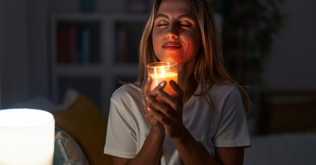 A woman holding a lit candle and smelling it while sitting on a bed with a lamp and pillows beside her.