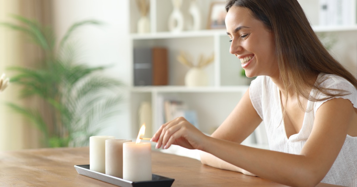 A female homeowner smiles as she lights a trio of candles on her dining room table. Each one is white.