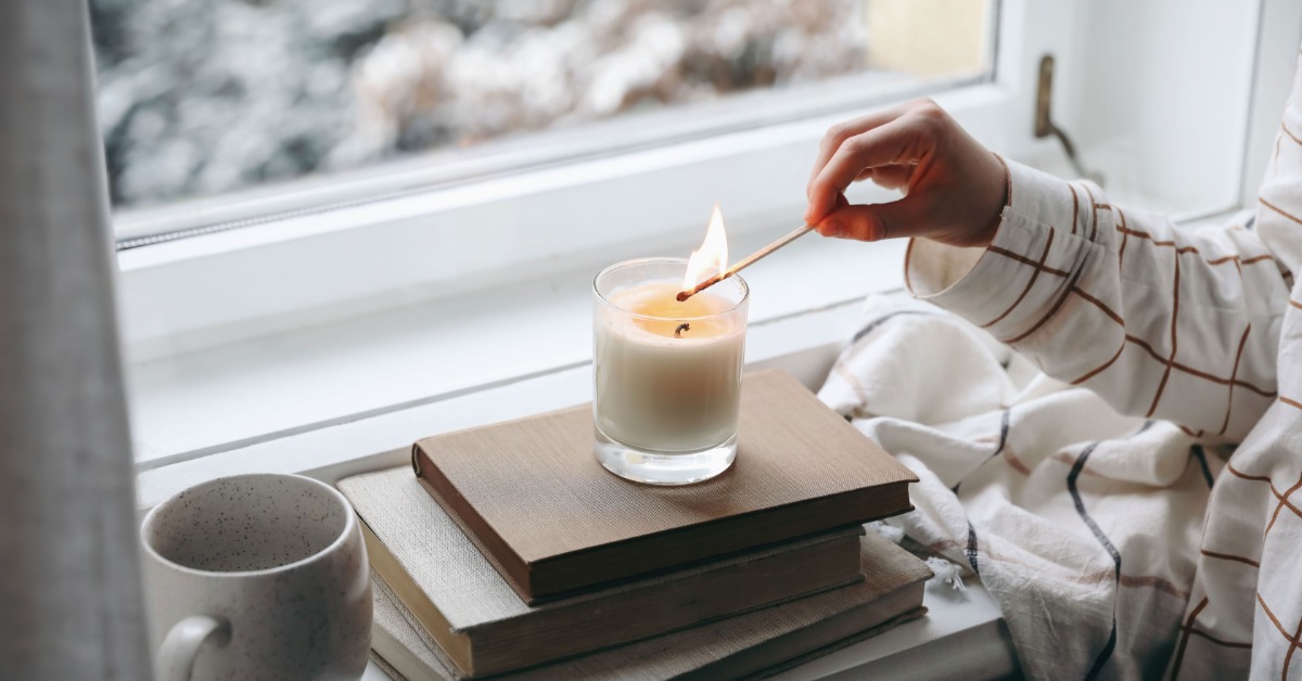 A small candle sits on a stack of books next to the bedroom window. A person lights the candle with a long match.