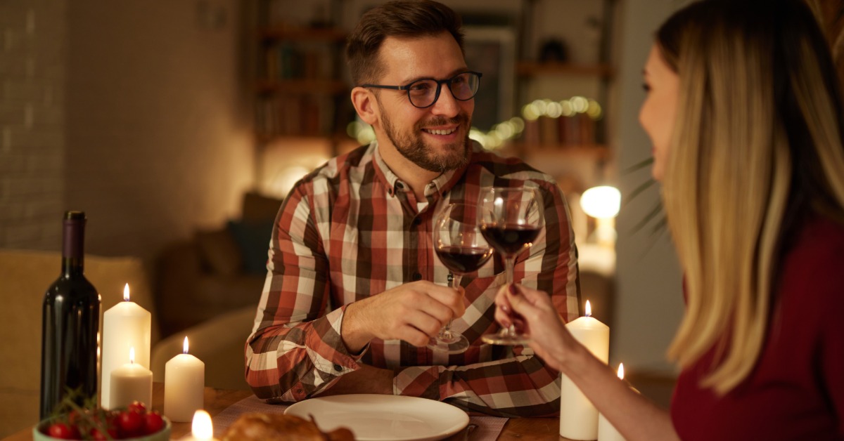 A happy couple at home sits at the dining room table and toasts with wine glasses. They have candles lit on the table.