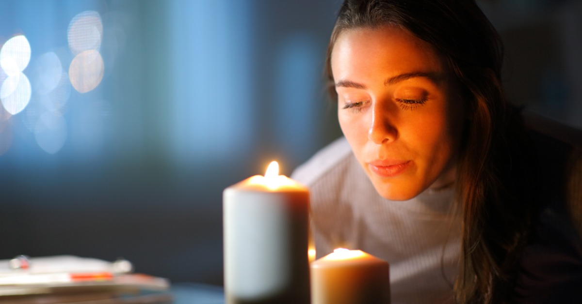 A young woman at home blowing out the candles on the table. She has two pillar candles lit in a dark space.