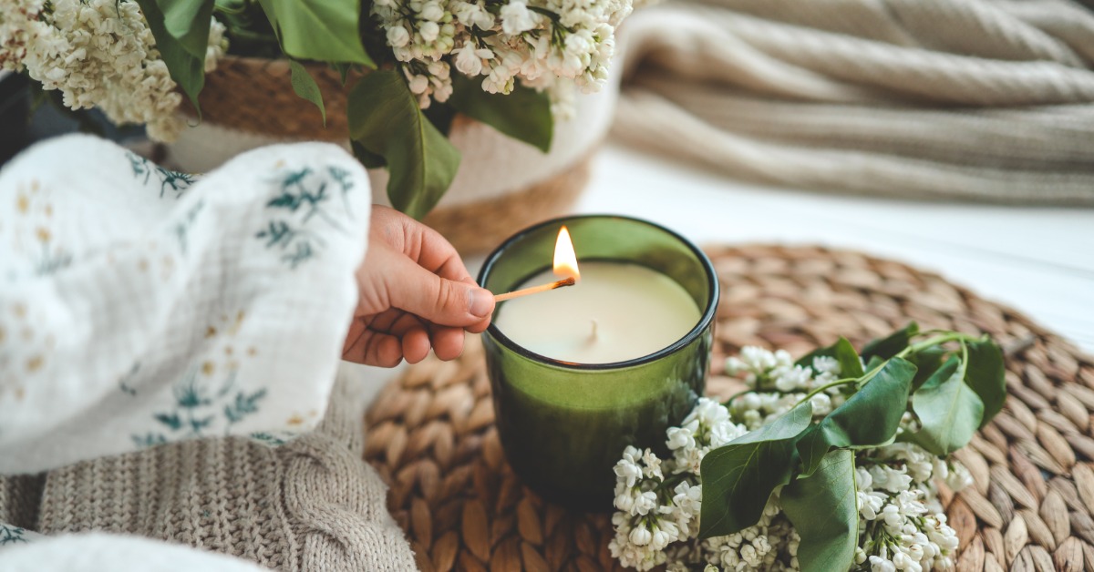 A close-up of someone lighting a green candle with a match in their left hand. There are white flowers near the candle.