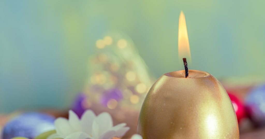 A lit golden egg-shaped candle stands beside a small nest of green eggs and white flowers, with colorful foil behind it.