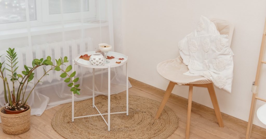 A white chair with flat pillows on it sitting near a table holding a few candles and some autumn leaves.