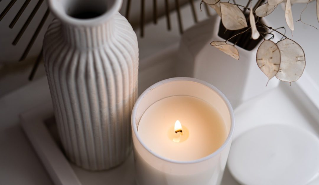 A close-up of a decorative tray on a table. The tray contains a burning candles, a vase, and the candle lid.