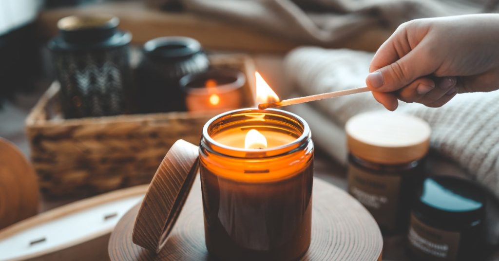 A close-up of someone lighting a candle with a long match. The candle burns with a lid leaning against it.