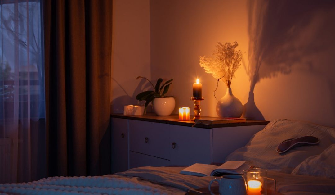 A cozy bedroom lit by candlelight at nighttime. There are two candles burning on the bedside table with a vase.