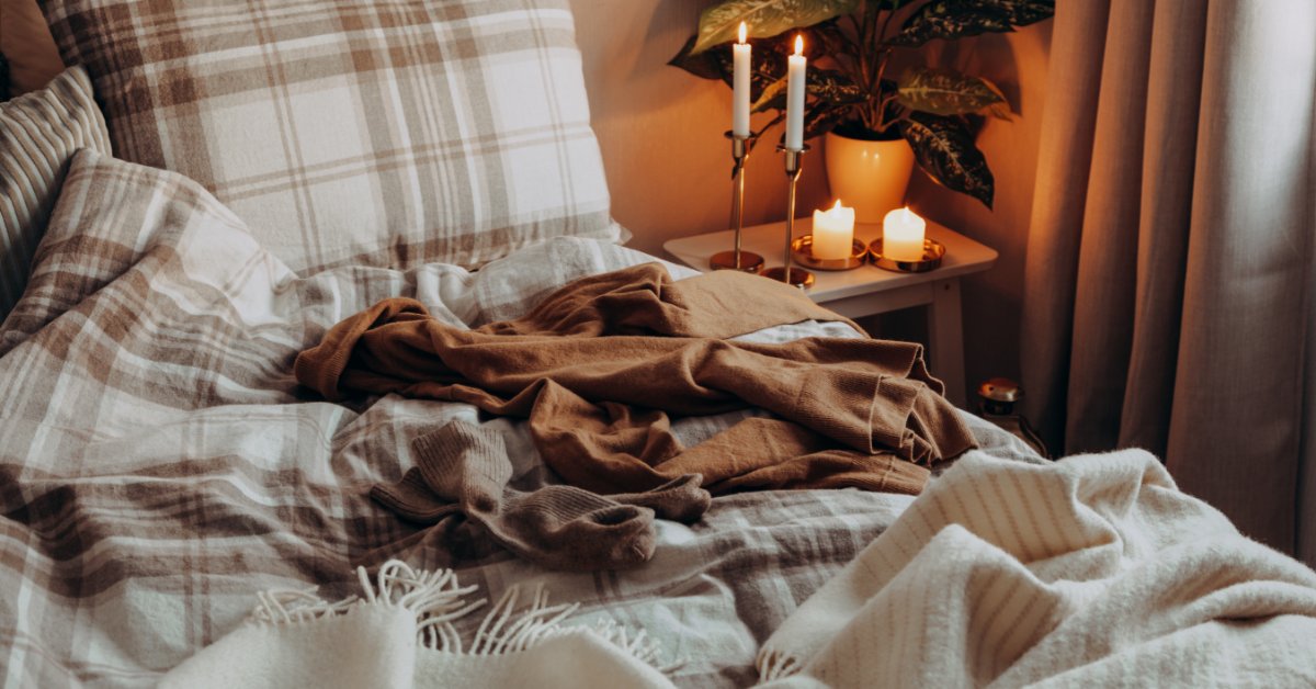 A messy bed in the bedroom with a candles burning on the righthand side nightstand. Two taper candles and two votives burn.