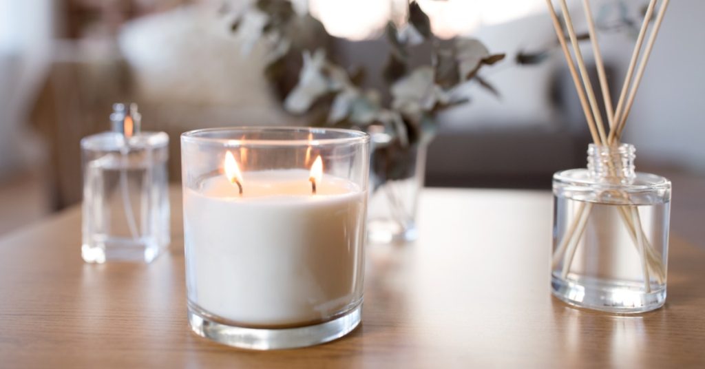 A small, two-wick votive glass candle burns on the table next to an oil reed diffuser. A bottle of perfume sits close.