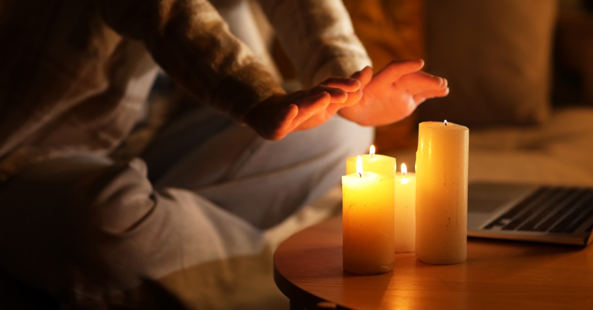 A person sits cozy on their couch at home as they place their hands over multiple lit pillar candles.