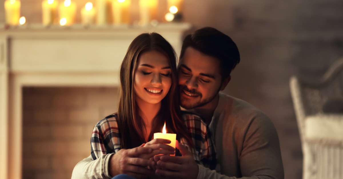 A couple sits cuddled together as they smile at a lit pillar candle in their hands. Candles line the mantle behind them.