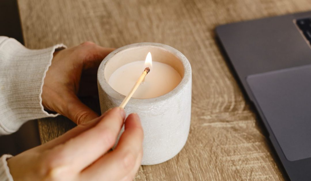 A person sits at the dining room table in front of a laptop as they light a small candle with a match.