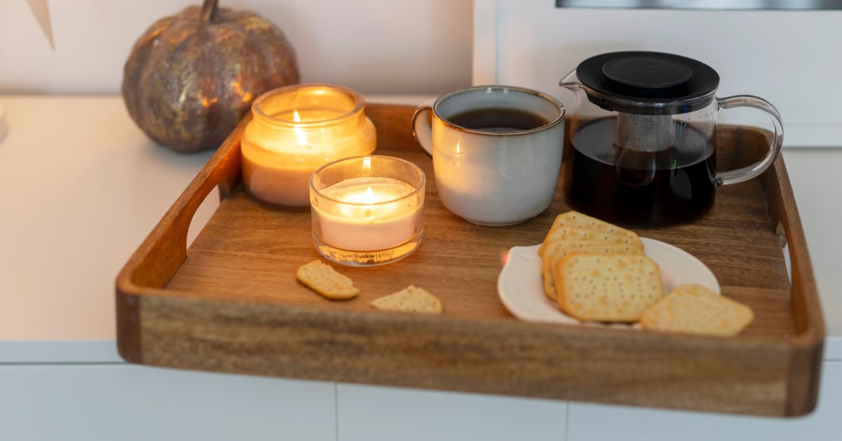 A wood tray with candles burning next to a teapot and a plate of crackers. There's a cup of tea next to the teapot.