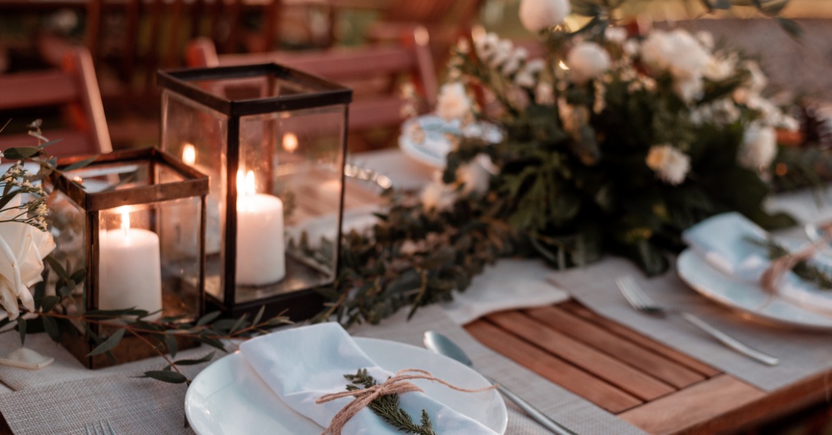 An outdoor dining table set with white china and rolled silverware. There are pillar candles in cages lit on the table.