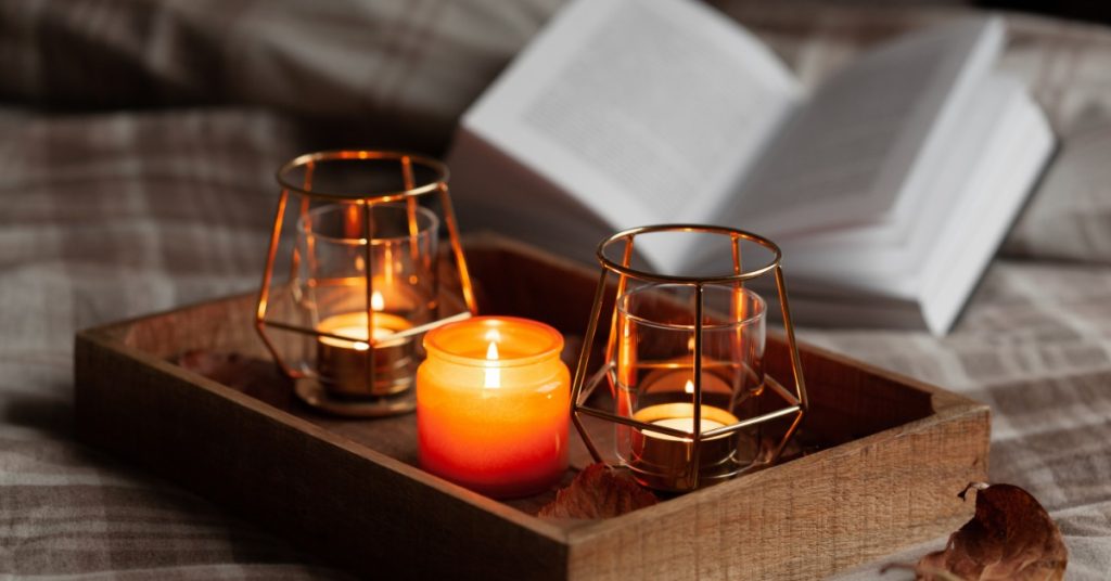 A wooden tray sits on a bed with three candles burning. There's an open book behind the tray to be read.